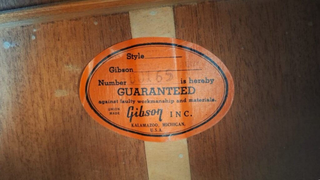 Auf einem orangefarbenen, ovalen Gibson-Etikett im Inneren des Holzkörpers der Gibson SJN 1968 Akustikgitarre steht „GUARANTEED against faulty workmanship and materials“ (Garantie gegen Verarbeitungs- und Materialfehler), darunter ist die handgeschriebene Nummer 7651 und der Aufdruck „Gibson Inc., Kalamazoo, Michigan, USA“ zu sehen. Auf einem orangefarbenen, ovalen Gibson-Etikett im Inneren des Holzkörpers der Gibson SJN 1968 Akustikgitarre steht „GUARANTEED against faulty workmanship and materials“ (Garantie gegen Verarbeitungs- und Materialfehler), darunter ist die handgeschriebene Nummer 7651 und der Aufdruck „Gibson Inc., Kalamazoo, Michigan, USA“ zu sehen.