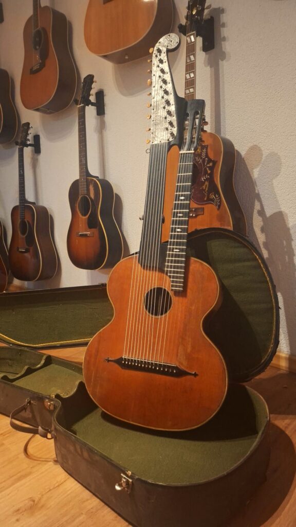 Eine Kontragitarre von Gustav Keller Stauffer im Stil von 1900 ist in einem offenen, grün ausgekleideten Kasten ausgestellt, umgeben von verschiedenen Akustikgitarren, die an der Wand eines Raums mit Holzboden hängen. Eine Kontragitarre von Gustav Keller Stauffer im Stil von 1900 ist in einem offenen, grün ausgekleideten Kasten ausgestellt, umgeben von verschiedenen Akustikgitarren, die an der Wand eines Raums mit Holzboden hängen.