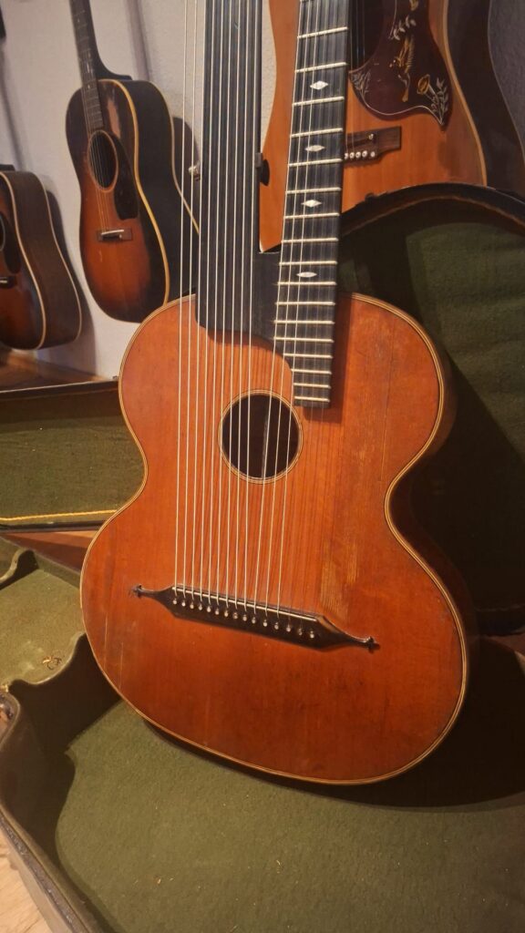 Eine Kontragitarre von Gustav Keller Stauffer im Stil von 1900 ist in einem offenen grünen Koffer ausgestellt, umgeben von anderen Akustikgitarren, die im Hintergrund an einer Wand hängen. Eine Kontragitarre von Gustav Keller Stauffer im Stil von 1900 ist in einem offenen grünen Koffer ausgestellt, umgeben von anderen Akustikgitarren, die im Hintergrund an einer Wand hängen.