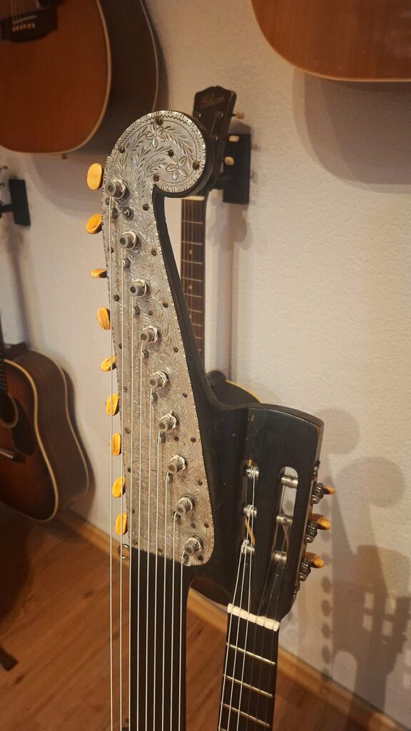 Eine Nahaufnahme des kunstvoll verzierten Metallkopfes der Gustav Keller Stauffer Stil 1900 Kontragitarre mit eingravierten Blumenmotiven und orangefarbenen Stimmwirbeln, umgeben von weiteren an einer Wand hängenden Akustikgitarren. Eine Nahaufnahme des kunstvoll verzierten Metallkopfes der Gustav Keller Stauffer Stil 1900 Kontragitarre mit eingravierten Blumenmotiven und orangefarbenen Stimmwirbeln, umgeben von weiteren an einer Wand hängenden Akustikgitarren.