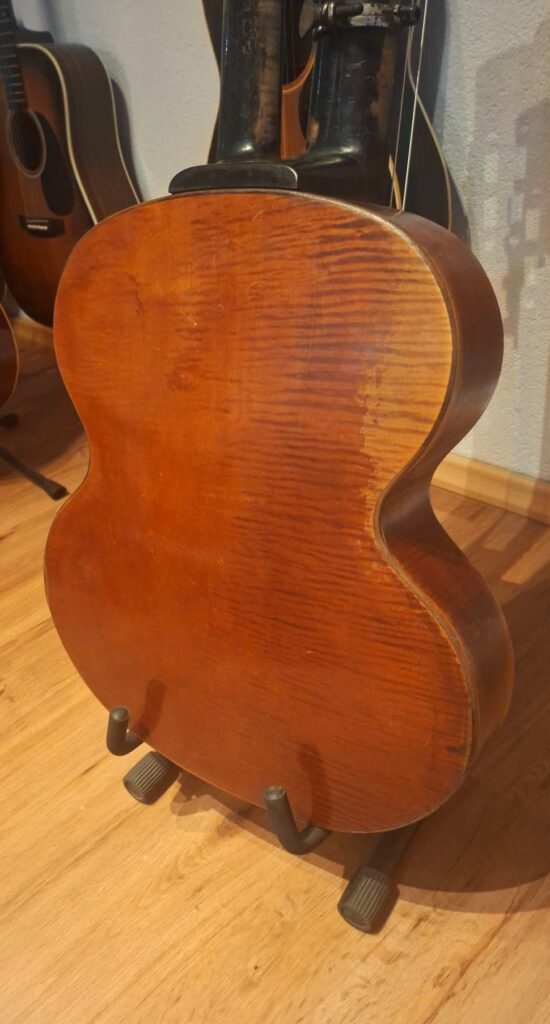 Eine Kontragitarre von Gustav Keller Stauffer im Stil von 1900 mit einer satten braunen Lackierung steht aufrecht auf einem Gitarrenständer auf einem Holzboden, im Hintergrund sind teilweise weitere Gitarren zu sehen. Eine Kontragitarre von Gustav Keller Stauffer im Stil von 1900 mit einer satten braunen Lackierung steht aufrecht auf einem Gitarrenständer auf einem Holzboden, im Hintergrund sind teilweise weitere Gitarren zu sehen.