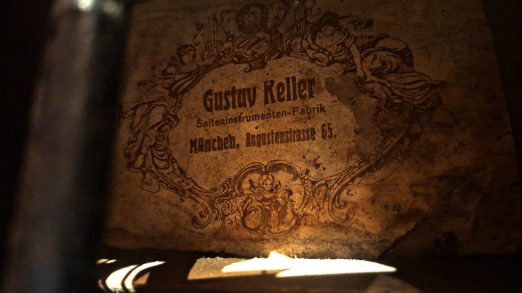 Auf einem Vintage-Etikett der „Gustav Keller Stauffer Stil 1900 Kontragitarre“ ist die Aufschrift „Gustav Keller Saiteninstrumenten-Fabrik, München, Augustenstraße 65“ zu sehen, begleitet von kunstvollen Illustrationen auf gealtertem Papier, das vom Sonnenlicht beleuchtet wird. Auf einem Vintage-Etikett der „Gustav Keller Stauffer Stil 1900 Kontragitarre“ ist die Aufschrift „Gustav Keller Saiteninstrumenten-Fabrik, München, Augustenstraße 65“ zu sehen, begleitet von kunstvollen Illustrationen auf gealtertem Papier, das vom Sonnenlicht beleuchtet wird.