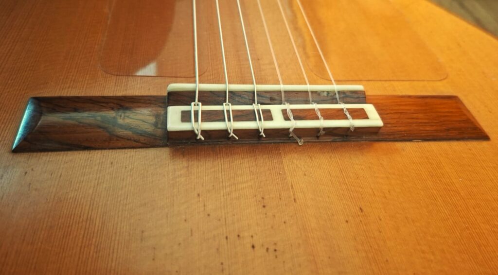 Nahaufnahme des Stegs einer „Negra Flamenco“-Gitarre von Juan Lopez Aguilarte aus dem Jahr 1984, die die detaillierte Holzmaserung und die am Steg befestigten Nylonsaiten zeigt, wobei ein Teil der Decke der Gitarre zu sehen ist. Warme Beleuchtung bringt die natürlichen Holzfarben und -strukturen besonders gut zur Geltung. Nahaufnahme des Stegs einer „Negra Flamenco“-Gitarre von Juan Lopez Aguilarte aus dem Jahr 1984, die die detaillierte Holzmaserung und die am Steg befestigten Nylonsaiten zeigt, wobei ein Teil der Decke der Gitarre zu sehen ist. Warme Beleuchtung bringt die natürlichen Holzfarben und -strukturen besonders gut zur Geltung.