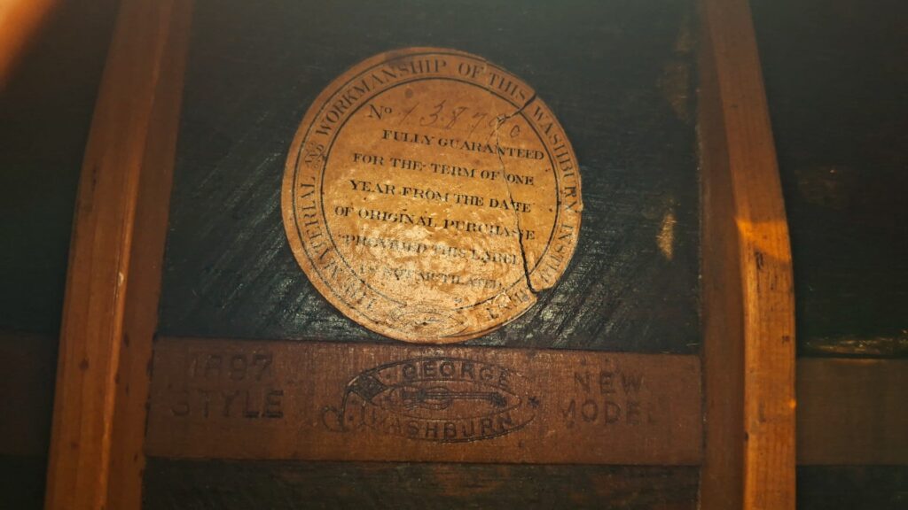 Nahaufnahme des Vintage-Etiketts der Washburn 1900 Parlor Akustikgitarre im Inneren ihres dunklen, holzgemaserten Korpus, auf dem Seriennummern, Garantieinformationen und der Name „Geo. Washburn“ sowie eingeprägte Markierungen zu sehen sind. Nahaufnahme des Vintage-Etiketts der Washburn 1900 Parlor Akustikgitarre im Inneren ihres dunklen, holzgemaserten Korpus, auf dem Seriennummern, Garantieinformationen und der Name „Geo. Washburn“ sowie eingeprägte Markierungen zu sehen sind.