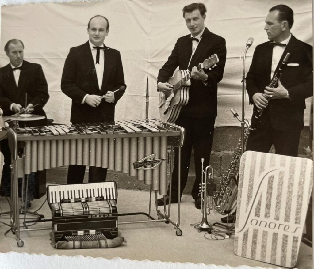 Vier Männer in Smokings spielen vor einem weißen Hintergrund verschiedene Instrumente – Vibraphon, Akkordeon, die Artur-Lang-Jazzgitarre aus den 1950er Jahren und Klarinette. Neben ihnen ist ein Schild mit der Aufschrift „Sonores“ und Noten zu sehen. Vier Männer in Smokings spielen vor einem weißen Hintergrund verschiedene Instrumente – Vibraphon, Akkordeon, die Artur-Lang-Jazzgitarre aus den 1950er Jahren und Klarinette. Neben ihnen ist ein Schild mit der Aufschrift „Sonores“ und Noten zu sehen.