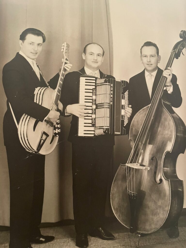 Drei Männer in Anzügen posieren mit Musikinstrumenten – einer elektrischen Jazzgitarre von Artur Lang aus den 1950er Jahren, einem Akkordeon und einem Kontrabass – vor einem Vorhang. Das Bild ist in Schwarz-Weiß gehalten. Drei Männer in Anzügen posieren mit Musikinstrumenten – einer elektrischen Jazzgitarre von Artur Lang aus den 1950er Jahren, einem Akkordeon und einem Kontrabass – vor einem Vorhang. Das Bild ist in Schwarz-Weiß gehalten.