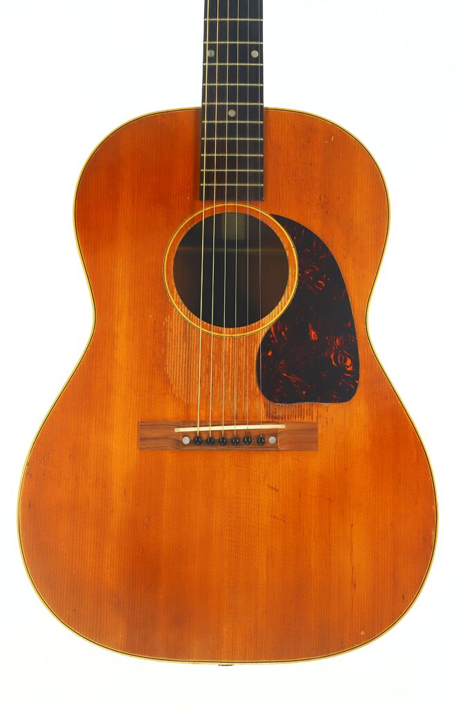 Eine Gibson LG-3 Akustikgitarre aus dem Jahr 1954 mit naturbelassener Holzoberfläche, schwarzem Schlagbrett, Stahlsaiten und sichtbaren Gebrauchsspuren am Korpus ist vor einem schlichten weißen Hintergrund abgebildet. Eine Gibson LG-3 Akustikgitarre aus dem Jahr 1954 mit naturbelassener Holzoberfläche, schwarzem Schlagbrett, Stahlsaiten und sichtbaren Gebrauchsspuren am Korpus ist vor einem schlichten weißen Hintergrund abgebildet.