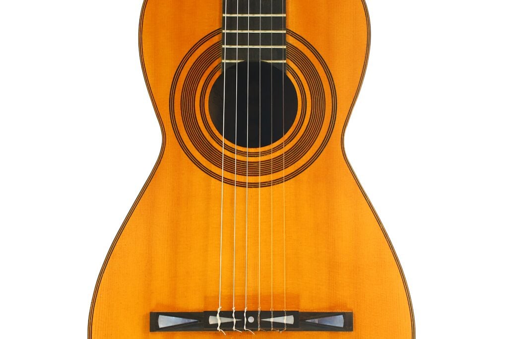 Eine Nahaufnahme der Antonio Garcia de Lorca 1847 Replika Klassische Gitarre, die den Holzkorpus, die sechs Saiten, das Schallloch mit kreisförmigen Verzierungen und einen Teil des Stegs vor einem weißen Hintergrund zeigt. Eine Nahaufnahme der Antonio Garcia de Lorca 1847 Replika Klassische Gitarre, die den Holzkorpus, die sechs Saiten, das Schallloch mit kreisförmigen Verzierungen und einen Teil des Stegs vor einem weißen Hintergrund zeigt.