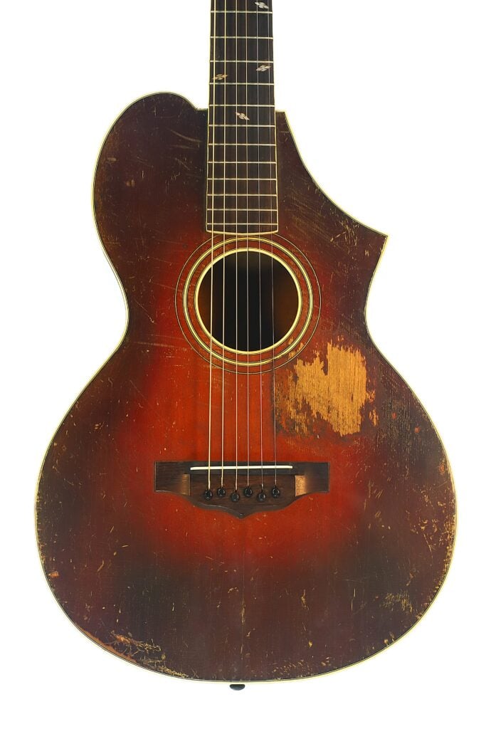 Eine Nahaufnahme einer abgenutzten Vintage-Akustikgitarre vom Typ „Epiphone Recording B 1931“ mit einem rotbraunen Korpus, sichtbaren Kratzern und einer großen Stelle an der rechten Seite, an der der Lack fehlt. Eine Nahaufnahme einer abgenutzten Vintage-Akustikgitarre vom Typ „Epiphone Recording B 1931“ mit einem rotbraunen Korpus, sichtbaren Kratzern und einer großen Stelle an der rechten Seite, an der der Lack fehlt.