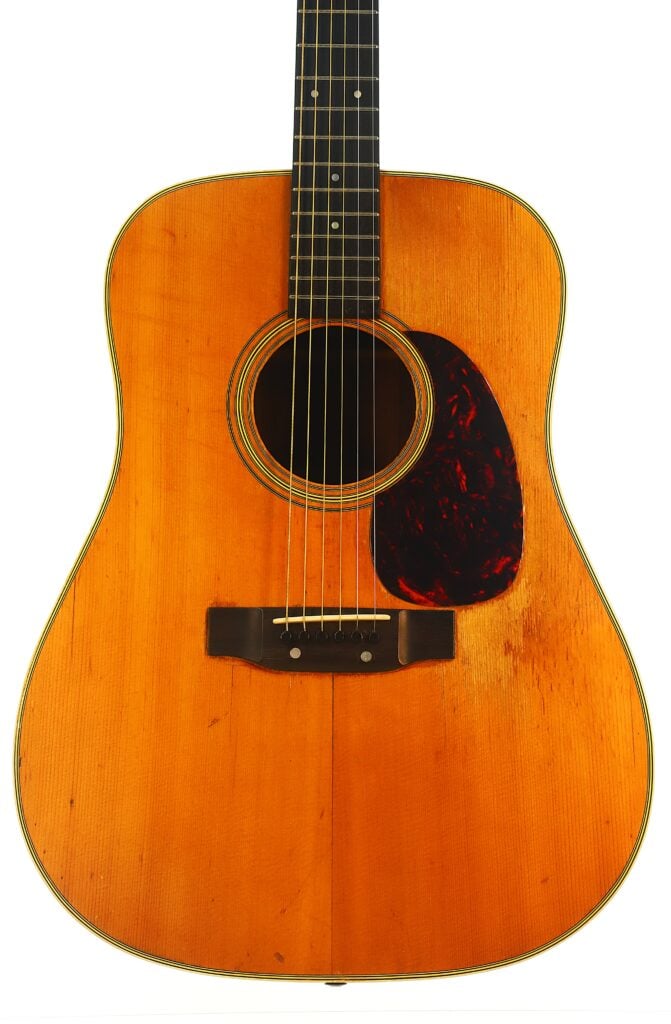Eine Nahaufnahme einer Martin D-28 Akustikgitarre aus dem Jahr 1965, die den Holzkorpus, die sichtbaren Saiten, das dunkle Schlagbrett und Gebrauchsspuren unterhalb der Saiten zeigt. Eine Nahaufnahme einer Martin D-28 Akustikgitarre aus dem Jahr 1965, die den Holzkorpus, die sichtbaren Saiten, das dunkle Schlagbrett und Gebrauchsspuren unterhalb der Saiten zeigt.