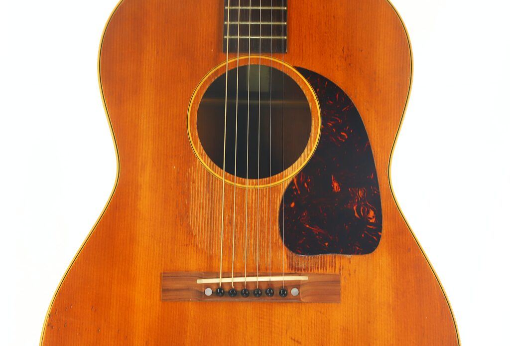 Nahaufnahme des Korpus einer Gibson LG-3 Akustikgitarre aus dem Jahr 1954 mit brauner Sunburst-Lackierung und sichtbarer Holzmaserung, einem dunklen Schlagbrett, sechs Saiten sowie deutlich erkennbarer Steg und Schallloch. Nahaufnahme des Korpus einer Gibson LG-3 Akustikgitarre aus dem Jahr 1954 mit brauner Sunburst-Lackierung und sichtbarer Holzmaserung, einem dunklen Schlagbrett, sechs Saiten sowie deutlich erkennbarer Steg und Schallloch.