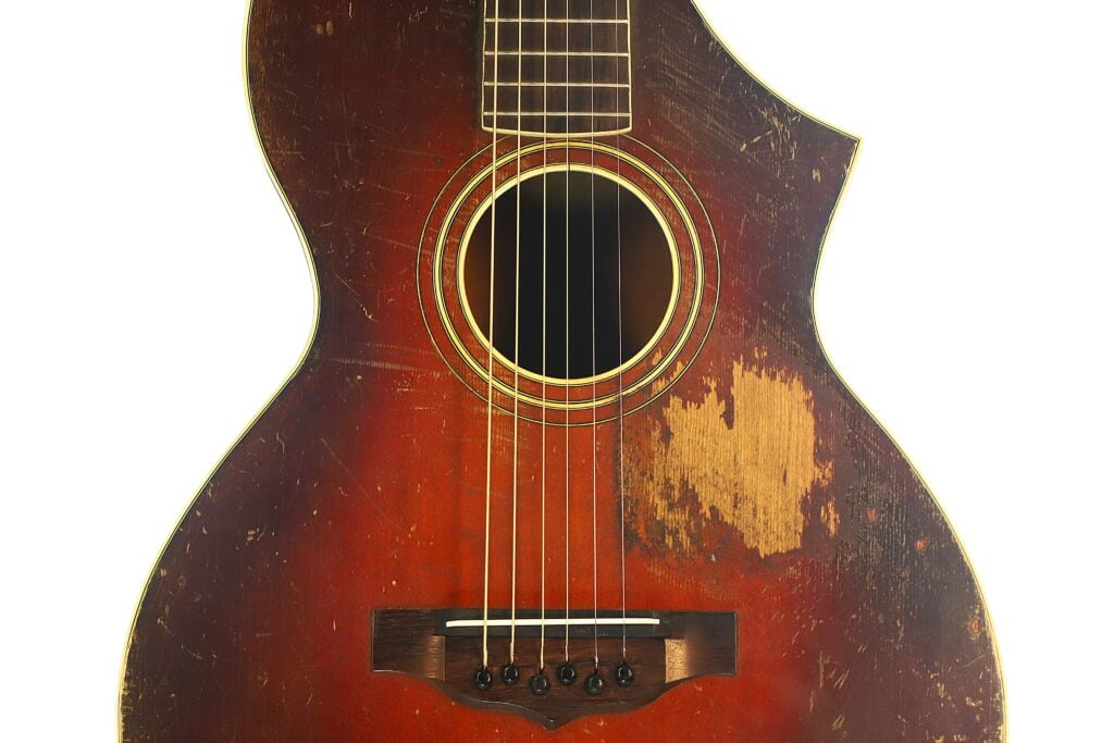 Nahaufnahme einer abgenutzten Vintage-Akustikgitarre vom Typ „Epiphone Recording B 1931“, die Kratzer und eine große Stelle mit fehlendem Lack am Korpus aufweist, wobei das gealterte Holz und die Metallsaiten vor einem weißen Hintergrund besonders hervorstechen. Nahaufnahme einer abgenutzten Vintage-Akustikgitarre vom Typ „Epiphone Recording B 1931“, die Kratzer und eine große Stelle mit fehlendem Lack am Korpus aufweist, wobei das gealterte Holz und die Metallsaiten vor einem weißen Hintergrund besonders hervorstechen.