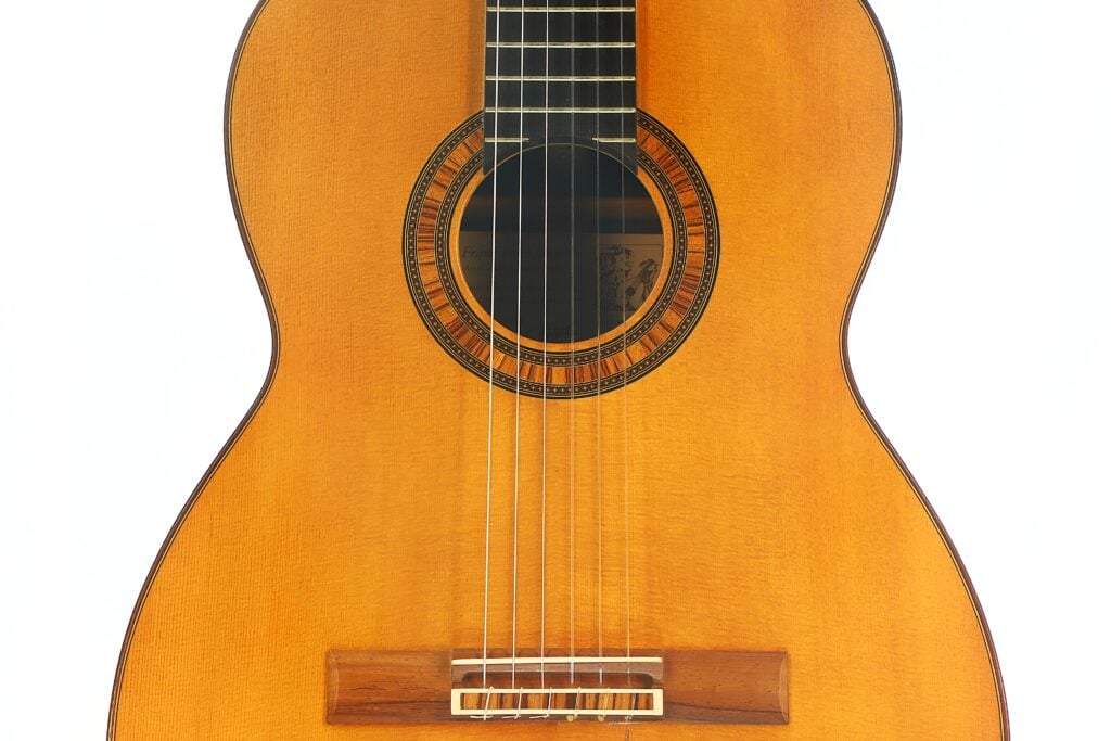 Nahaufnahme der Vorderseite der „Tosca Klassische Gitarre“ von Frank-Peter Dietrich aus dem Jahr 1998, zu sehen sind das Schallloch, die Zierrosette, die Saiten und ein Teil des Holzstegs vor weißem Hintergrund. Nahaufnahme der Vorderseite der „Tosca Klassische Gitarre“ von Frank-Peter Dietrich aus dem Jahr 1998, zu sehen sind das Schallloch, die Zierrosette, die Saiten und ein Teil des Holzstegs vor weißem Hintergrund.