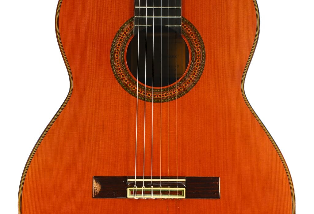 Nahaufnahme des Korpus einer Paulino Bernabe M-5 2008 Klassische Gitarre, die die Holzoberfläche, das Schallloch mit der Zierrosette, die Nylonsaiten und den Steg hervorhebt. Nahaufnahme des Korpus einer Paulino Bernabe M-5 2008 Klassische Gitarre, die die Holzoberfläche, das Schallloch mit der Zierrosette, die Nylonsaiten und den Steg hervorhebt.