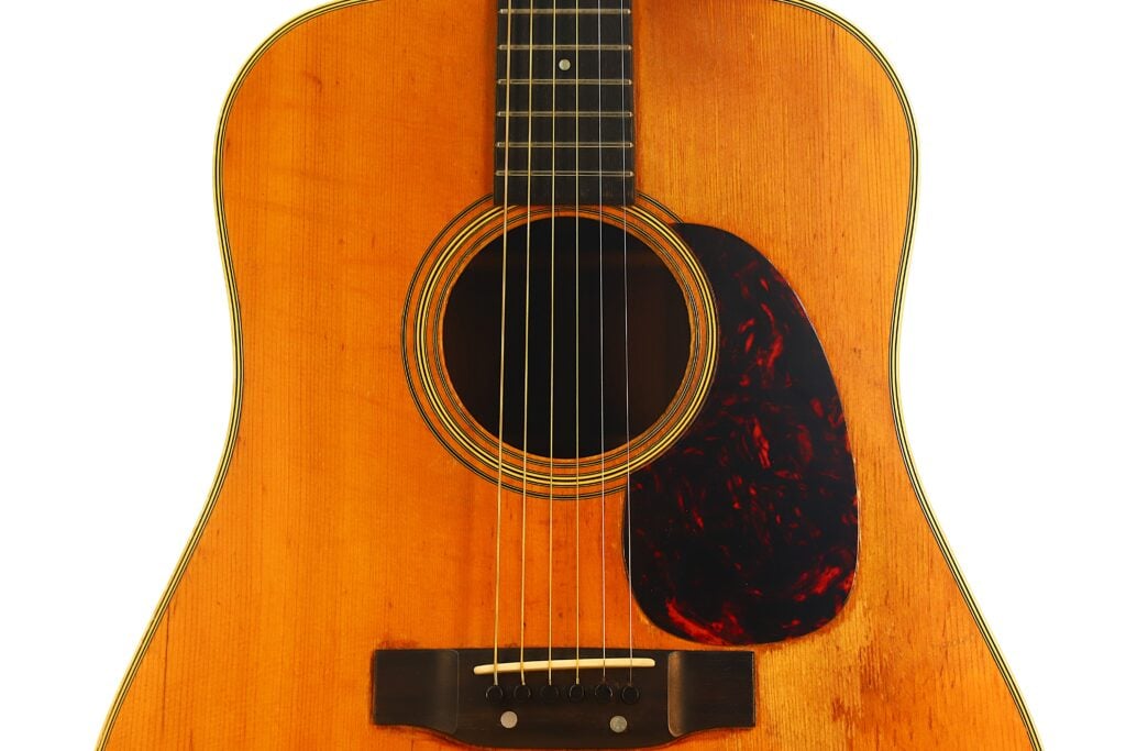 Nahaufnahme des Korpus der Martin D-28 Akustikgitarre aus dem Jahr 1965, die das Schallloch, die Saiten, das Schlagbrett und den Steg vor einem weißen Hintergrund zeigt. Nahaufnahme des Korpus der Martin D-28 Akustikgitarre aus dem Jahr 1965, die das Schallloch, die Saiten, das Schlagbrett und den Steg vor einem weißen Hintergrund zeigt.