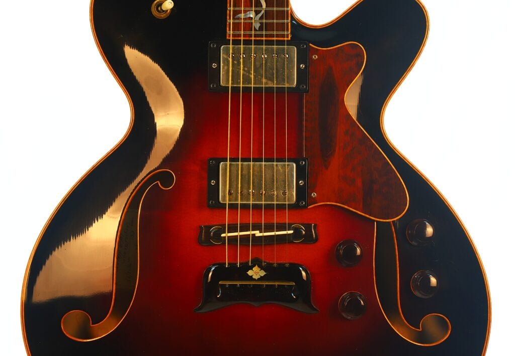 Nahaufnahme der DeLaHaye 1995 Elektro-Akustik-Gitarre, bei der der halbhohle Korpus mit Sunburst-Lackierung in Orange-, Rot- und Schwarztönen, die sichtbaren Saiten, Tonabnehmer, F-Löcher, Regler sowie die glänzende Oberfläche besonders hervorgehoben werden. Nahaufnahme der DeLaHaye 1995 Elektro-Akustik-Gitarre, bei der der halbhohle Korpus mit Sunburst-Lackierung in Orange-, Rot- und Schwarztönen, die sichtbaren Saiten, Tonabnehmer, F-Löcher, Regler sowie die glänzende Oberfläche besonders hervorgehoben werden.