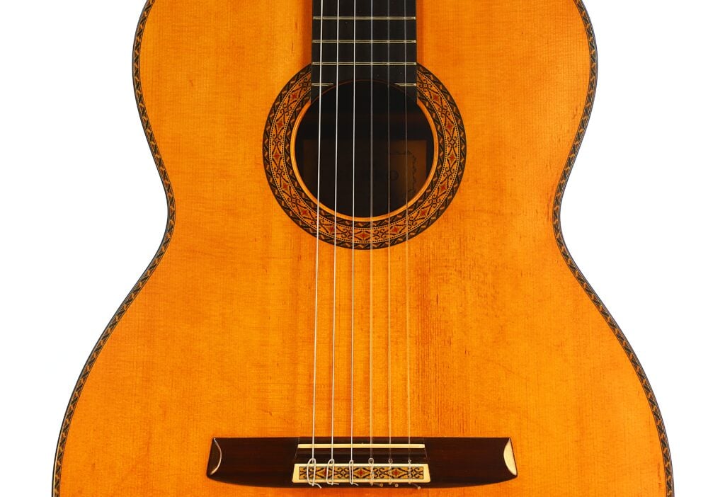 Nahaufnahme des Korpus einer klassischen Gitarre vom Typ „Masaru Kohno Modell 50“ aus dem Jahr 1979, auf der das Schallloch mit dekorativer Rosette, die sechs Saiten, der Holzsteg und die warmbraune Lackierung zu sehen sind. Nahaufnahme des Korpus einer klassischen Gitarre vom Typ „Masaru Kohno Modell 50“ aus dem Jahr 1979, auf der das Schallloch mit dekorativer Rosette, die sechs Saiten, der Holzsteg und die warmbraune Lackierung zu sehen sind.