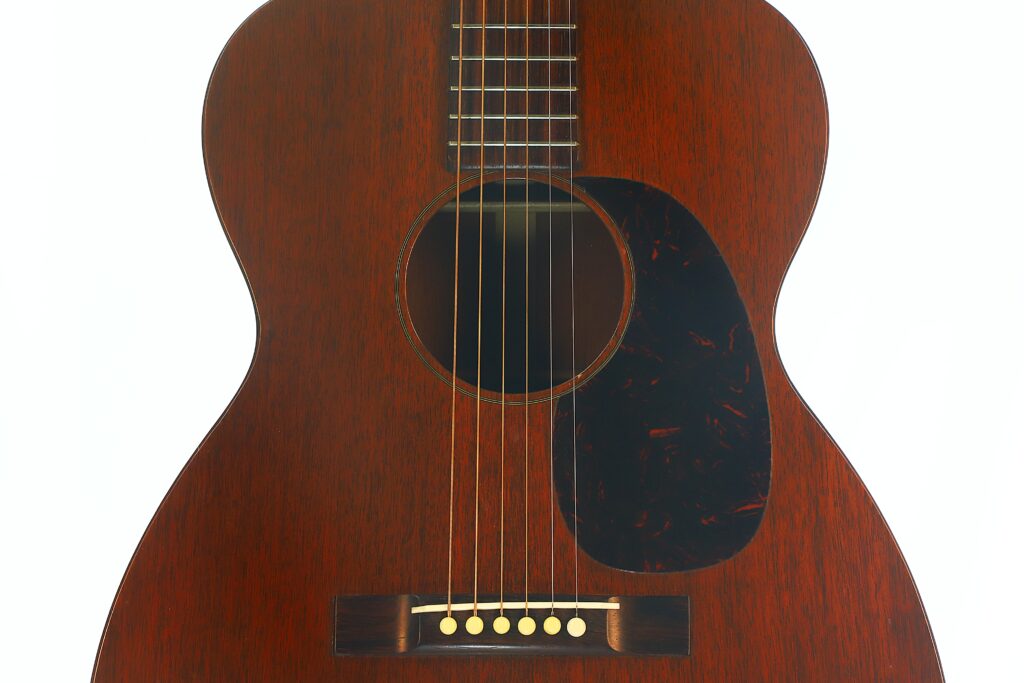 Nahaufnahme des Korpus einer Martin 00-17 Akustikgitarre aus dem Jahr 1941 mit dunkler Holzoberfläche, sechs Saiten, einem schwarzen Schlagbrett und einem Steg mit sechs Stiften. Der Hintergrund ist weiß. Nahaufnahme des Korpus einer Martin 00-17 Akustikgitarre aus dem Jahr 1941 mit dunkler Holzoberfläche, sechs Saiten, einem schwarzen Schlagbrett und einem Steg mit sechs Stiften. Der Hintergrund ist weiß.