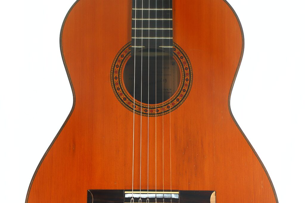 Eine Nahaufnahme des Korpus und der Saiten der Flamenco-Gitarre „Casa Arcangel Fernandez 1970“, die die Holzoberfläche hervorhebt und den Fokus auf das Schallloch und die dekorative Rosette legt. Eine Nahaufnahme des Korpus und der Saiten der Flamenco-Gitarre „Casa Arcangel Fernandez 1970“, die die Holzoberfläche hervorhebt und den Fokus auf das Schallloch und die dekorative Rosette legt.