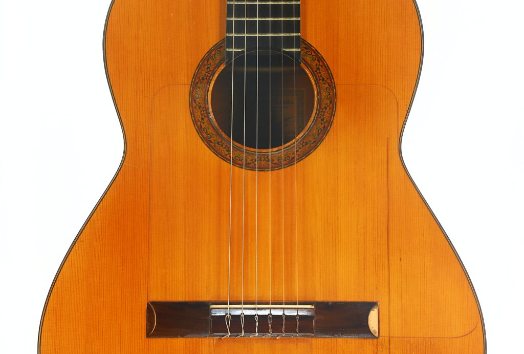 Nahaufnahme des Korpus einer Flamenco-Gitarre von Francisco Barba aus dem Jahr 1966 mit sechs Saiten, einer kunstvoll gearbeiteten Rosette, die das Schallloch umgibt, und einem Holzsteg vor einem schlichten weißen Hintergrund. Nahaufnahme des Korpus einer Flamenco-Gitarre von Francisco Barba aus dem Jahr 1966 mit sechs Saiten, einer kunstvoll gearbeiteten Rosette, die das Schallloch umgibt, und einem Holzsteg vor einem schlichten weißen Hintergrund.
