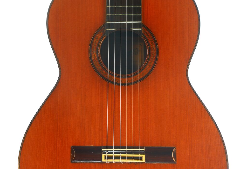 Nahaufnahme des Korpus und der Saiten einer Ramirez 1a 1985 Klassische Gitarre, mit Fokus auf die hölzerne Decke, die das Schallloch umgebende Zierrosette und den Steg. Nahaufnahme des Korpus und der Saiten einer Ramirez 1a 1985 Klassische Gitarre, mit Fokus auf die hölzerne Decke, die das Schallloch umgebende Zierrosette und den Steg.