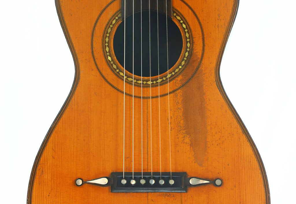 Nahaufnahme des Korpus einer „Romantikgitarre“ von Louis Panormo aus dem Jahr 1827 mit einem runden Schallloch, das von dekorativen Verzierungen umgeben ist, sowie sechs Saiten. Auf der rechten Seite des Holzes ist ein Fleck zu erkennen. Nahaufnahme des Korpus einer „Romantikgitarre“ von Louis Panormo aus dem Jahr 1827 mit einem runden Schallloch, das von dekorativen Verzierungen umgeben ist, sowie sechs Saiten. Auf der rechten Seite des Holzes ist ein Fleck zu erkennen.