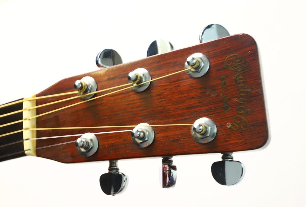 Nahaufnahme des Kopfes einer Martin D-28 Akustikgitarre aus dem Jahr 1965, auf der die Stimmwirbel, die Saiten und das „Martin & Co.“-Logo auf einer Holzoberfläche vor einem schlichten weißen Hintergrund zu sehen sind. Nahaufnahme des Kopfes einer Martin D-28 Akustikgitarre aus dem Jahr 1965, auf der die Stimmwirbel, die Saiten und das „Martin & Co.“-Logo auf einer Holzoberfläche vor einem schlichten weißen Hintergrund zu sehen sind.