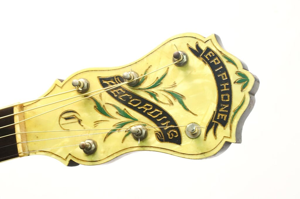 Die kunstvoll verzierte Kopfplatte einer Epiphone Recording B 1931 Akustikgitarre ist gelb lackiert und mit Blumenmotiven verziert. Die Aufschriften „Epiphone“ und „Recording“ sind in schwarzer Schreibschrift angebracht, und sechs Stimmwirbel sowie einige Gitarrensaiten sind zu sehen. Die kunstvoll verzierte Kopfplatte einer Epiphone Recording B 1931 Akustikgitarre ist gelb lackiert und mit Blumenmotiven verziert. Die Aufschriften „Epiphone“ und „Recording“ sind in schwarzer Schreibschrift angebracht, und sechs Stimmwirbel sowie einige Gitarrensaiten sind zu sehen.