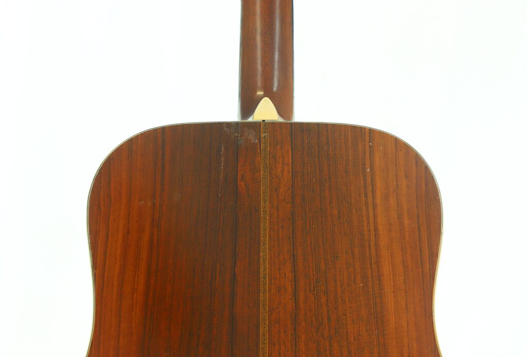 Nahaufnahme der Rückseite einer Martin D-28 1941 Pre-War Akustikgitarre, die die reichhaltige Holzmaserung und den dekorativen Mittelstreifen hervorhebt, der vertikal entlang des Korpus verläuft, mit einer hellen Halsfußkappe am Halsansatz. Nahaufnahme der Rückseite einer Martin D-28 1941 Pre-War Akustikgitarre, die die reichhaltige Holzmaserung und den dekorativen Mittelstreifen hervorhebt, der vertikal entlang des Korpus verläuft, mit einer hellen Halsfußkappe am Halsansatz.