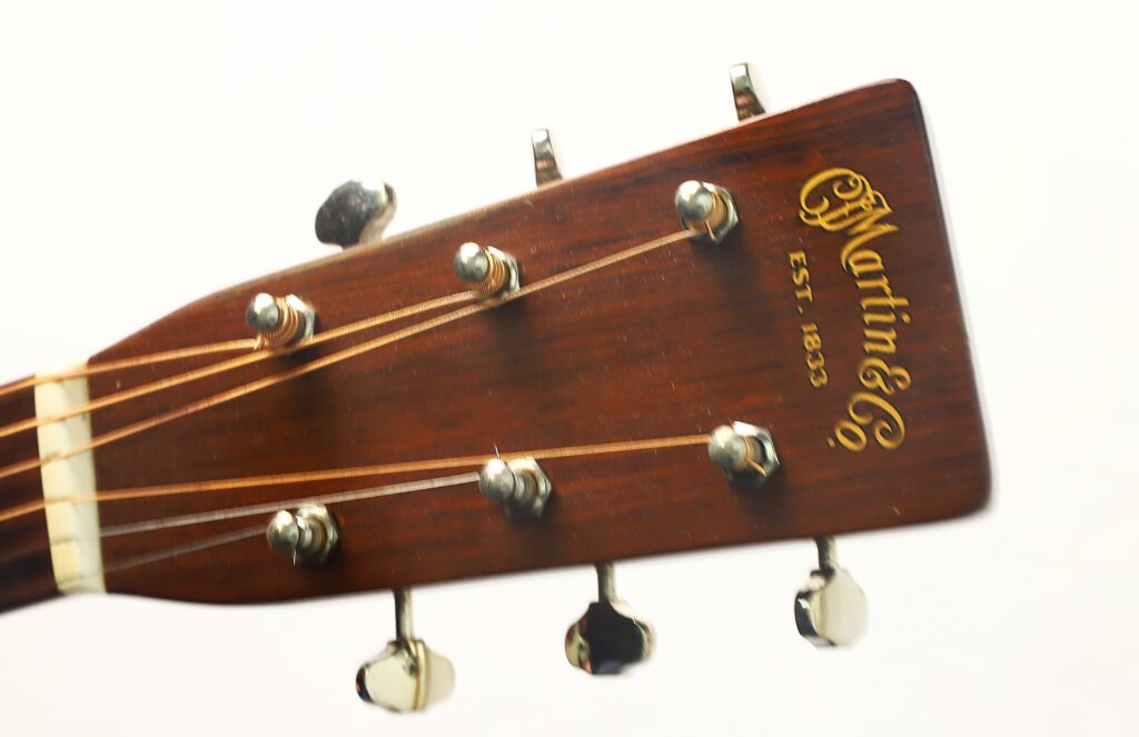 Nahaufnahme des Kopfes einer Martin 00-17 Akustikgitarre aus dem Jahr 1941, mit Fokus auf die Stimmwirbel, die Saiten und das Martin & Co-Logo mit der Aufschrift „EST. 1833“ auf der Holzoberfläche. Nahaufnahme des Kopfes einer Martin 00-17 Akustikgitarre aus dem Jahr 1941, mit Fokus auf die Stimmwirbel, die Saiten und das Martin & Co-Logo mit der Aufschrift „EST. 1833“ auf der Holzoberfläche.