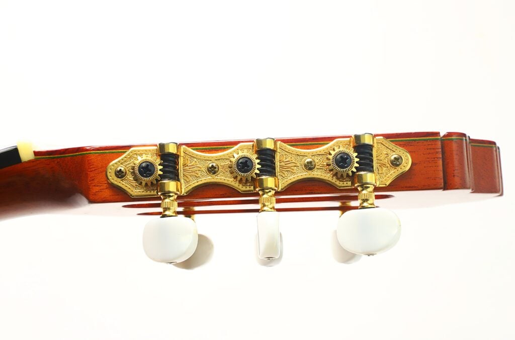 Nahaufnahme des Kopfes der Paulino Bernabe M-5 2008 Klassische Gitarre mit drei weißen Stimmwirbeln auf verzierten goldenen Stimmmechaniken vor einem hölzernen Hintergrund. Nahaufnahme des Kopfes der Paulino Bernabe M-5 2008 Klassische Gitarre mit drei weißen Stimmwirbeln auf verzierten goldenen Stimmmechaniken vor einem hölzernen Hintergrund.