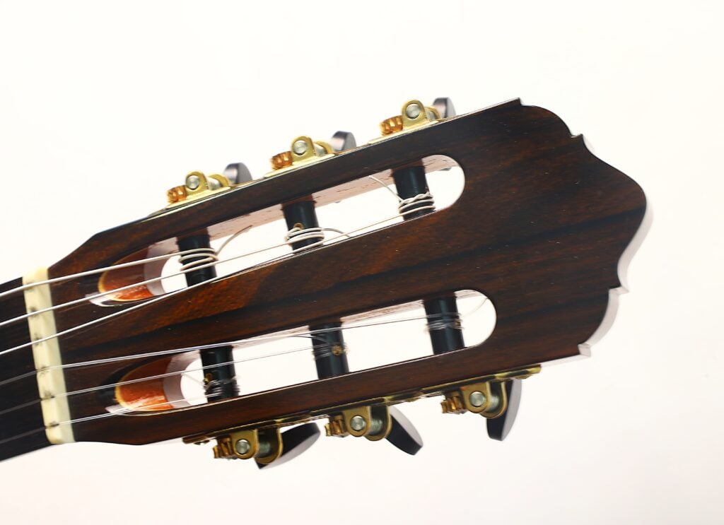 Nahaufnahme des Kopfstücks einer Lester DeVoe 2020 Negra Flamenco-Gitarre mit dunkler Holzoberfläche, goldenen Stimmwirbeln und Nylonsaiten vor einem schlichten weißen Hintergrund. Nahaufnahme des Kopfstücks einer Lester DeVoe 2020 Negra Flamenco-Gitarre mit dunkler Holzoberfläche, goldenen Stimmwirbeln und Nylonsaiten vor einem schlichten weißen Hintergrund.