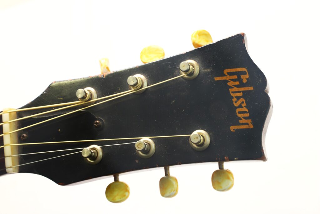 Nahaufnahme des Kopfes einer Gibson LG-3 Akustikgitarre aus dem Jahr 1954 mit sechs Stimmwirbeln und einem goldenen Gibson-Logo vor weißem Hintergrund. Nahaufnahme des Kopfes einer Gibson LG-3 Akustikgitarre aus dem Jahr 1954 mit sechs Stimmwirbeln und einem goldenen Gibson-Logo vor weißem Hintergrund.