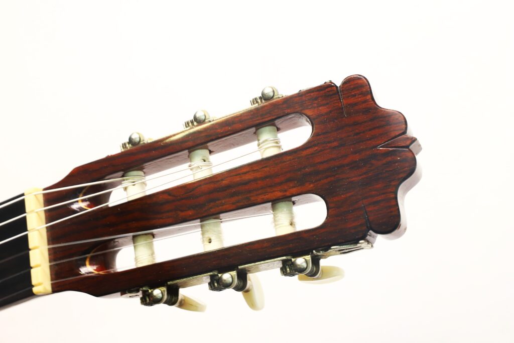 Nahaufnahme des Kopfstücks der Flamenco-Gitarre „Casa Arcangel Fernandez 1970“ mit hölzernen Stimmwirbeln, Saiten und einem hellen Hintergrund. Nahaufnahme des Kopfstücks der Flamenco-Gitarre „Casa Arcangel Fernandez 1970“ mit hölzernen Stimmwirbeln, Saiten und einem hellen Hintergrund.