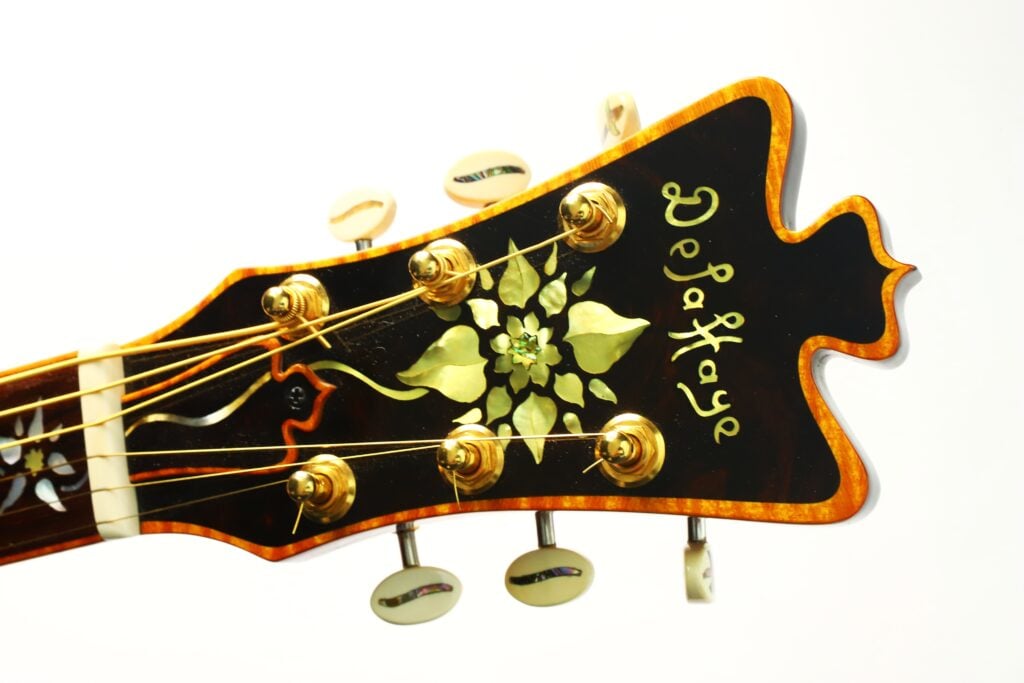 Eine Nahaufnahme des Kopfes der DeLaHaye Elektro-Akustik-Gitarre von 1995 zeigt goldene Stimmwirbel, florale Einlegearbeiten und den Schriftzug „DeLaHaye“ in kunstvoller Schrift auf dunklem Holzhintergrund. Eine Nahaufnahme des Kopfes der DeLaHaye Elektro-Akustik-Gitarre von 1995 zeigt goldene Stimmwirbel, florale Einlegearbeiten und den Schriftzug „DeLaHaye“ in kunstvoller Schrift auf dunklem Holzhintergrund.