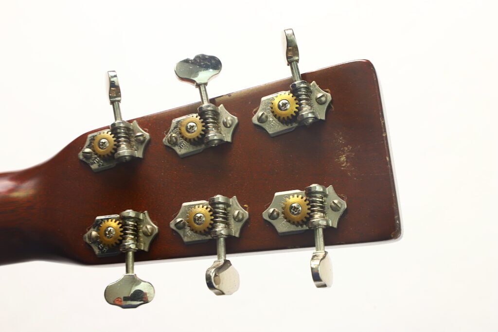 Nahaufnahme der Rückseite des Kopfes einer Martin 00-17 Akustikgitarre aus dem Jahr 1941, auf der sechs Metallstimmwirbel und Zahnräder vor einem schlichten weißen Hintergrund zu sehen sind. Nahaufnahme der Rückseite des Kopfes einer Martin 00-17 Akustikgitarre aus dem Jahr 1941, auf der sechs Metallstimmwirbel und Zahnräder vor einem schlichten weißen Hintergrund zu sehen sind.
