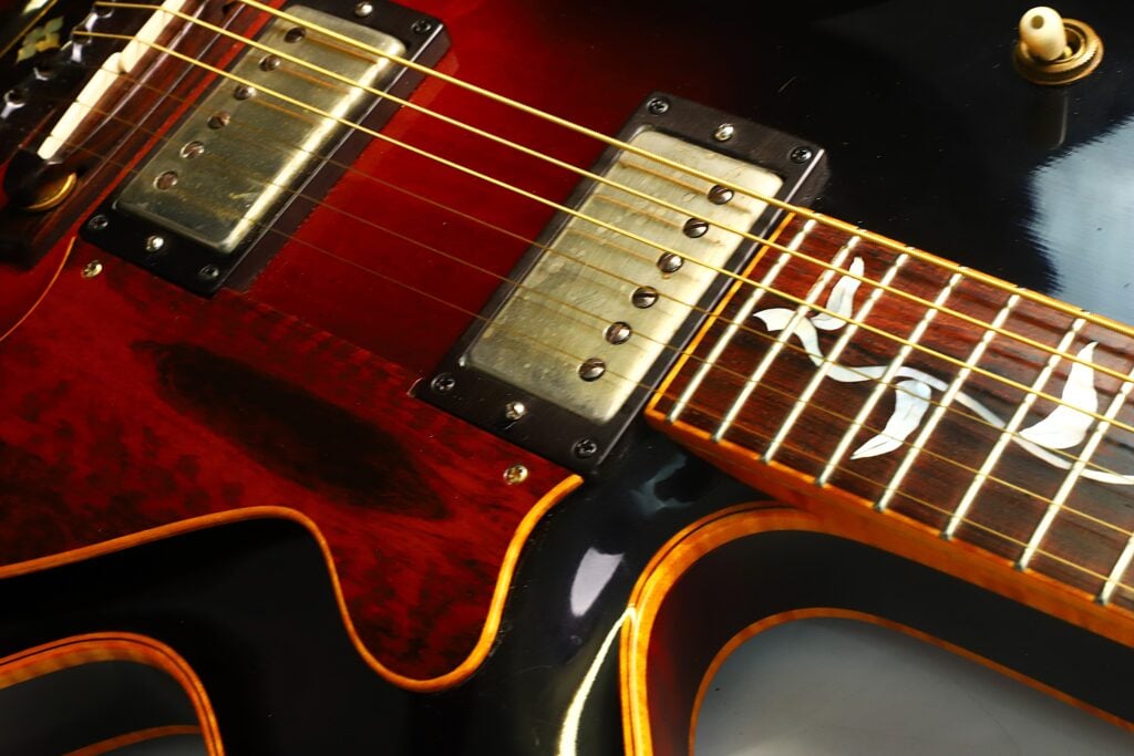 Nahaufnahme der DeLaHaye 1995 Elektro-Akustik-Gitarre, bei der die Saiten, Tonabnehmer, Regler und das dekorative Griffbrettdesign vor dem Hintergrund eines dunklen, glänzenden Korpus mit sichtbaren Holzmaserungsdetails besonders hervorgehoben werden. Nahaufnahme der DeLaHaye 1995 Elektro-Akustik-Gitarre, bei der die Saiten, Tonabnehmer, Regler und das dekorative Griffbrettdesign vor dem Hintergrund eines dunklen, glänzenden Korpus mit sichtbaren Holzmaserungsdetails besonders hervorgehoben werden.