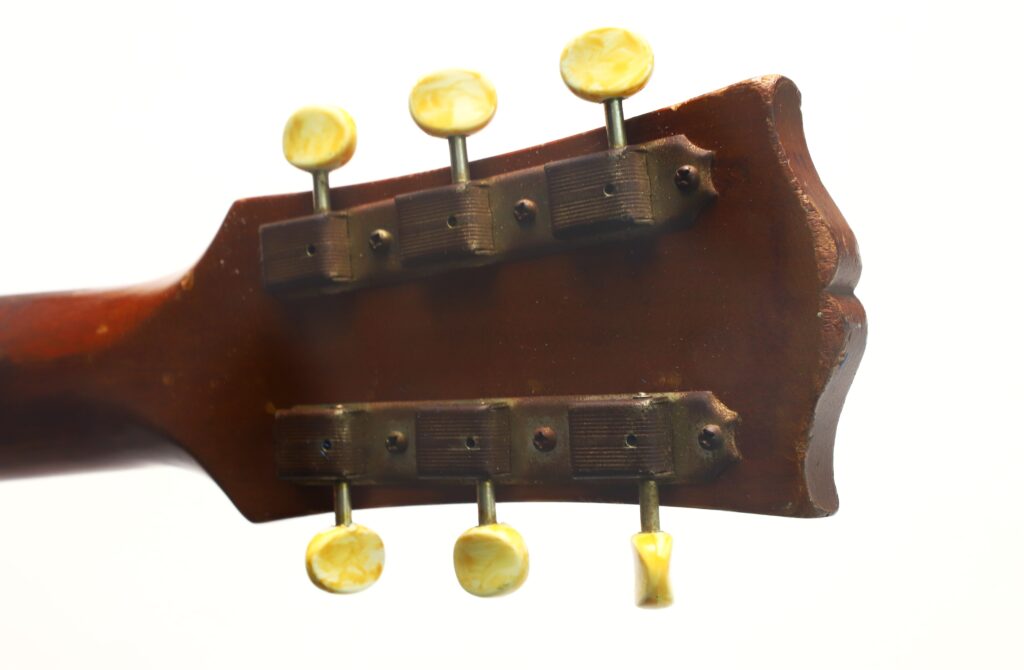 Nahaufnahme der Rückseite des Kopfes einer Gibson LG-3 Akustikgitarre aus dem Jahr 1954, auf der sechs gelbe Stimmwirbel und Metall-Stimmmechaniken vor weißem Hintergrund zu sehen sind. Nahaufnahme der Rückseite des Kopfes einer Gibson LG-3 Akustikgitarre aus dem Jahr 1954, auf der sechs gelbe Stimmwirbel und Metall-Stimmmechaniken vor weißem Hintergrund zu sehen sind.