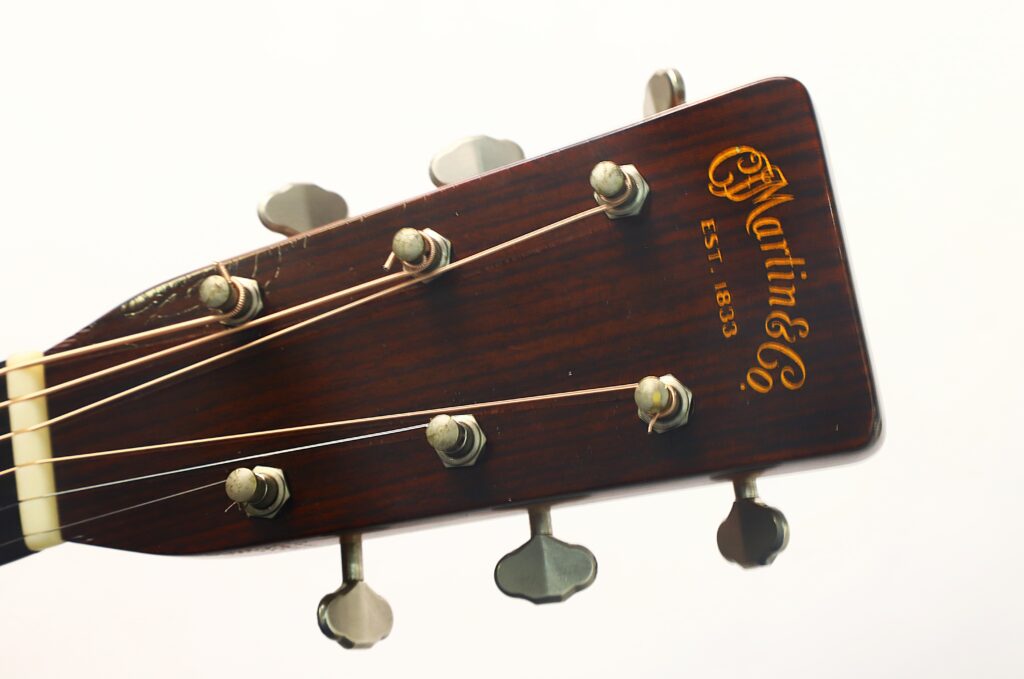 Nahaufnahme des Kopfes einer Martin D-28 1941 Pre-War Akustikgitarre, auf der die Stimmwirbel, die Saitenanordnung und das Martin & Co-Logo mit der Aufschrift „Est. 1833“ in goldener Schrift auf dunklem Holz zu sehen sind. Nahaufnahme des Kopfes einer Martin D-28 1941 Pre-War Akustikgitarre, auf der die Stimmwirbel, die Saitenanordnung und das Martin & Co-Logo mit der Aufschrift „Est. 1833“ in goldener Schrift auf dunklem Holz zu sehen sind.