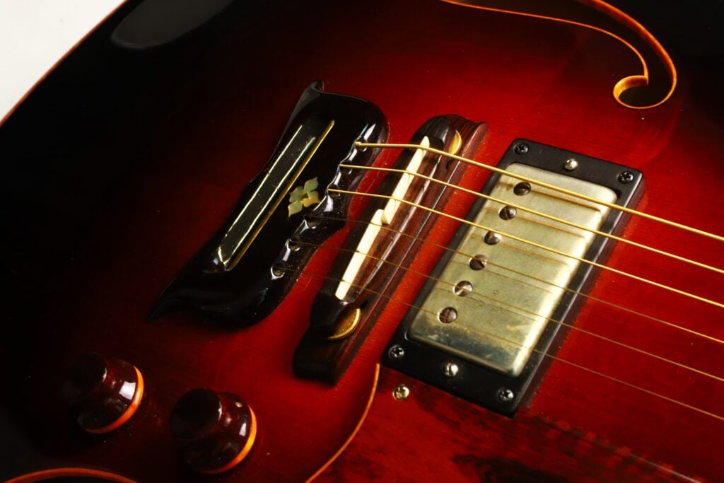 Nahaufnahme des Stegs, des Tonabnehmers und der Regler der DeLaHaye 1995 Elektro-Akustik-Gitarre auf einem glänzenden rot-schwarzen Korpus, die die Holzmaserung und die filigranen Metallkomponenten hervorhebt. Nahaufnahme des Stegs, des Tonabnehmers und der Regler der DeLaHaye 1995 Elektro-Akustik-Gitarre auf einem glänzenden rot-schwarzen Korpus, die die Holzmaserung und die filigranen Metallkomponenten hervorhebt.