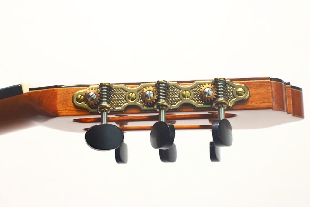 Nahaufnahme des Kopfes und der Stimmwirbel der Lester DeVoe 2020 Negra Flamenco-Gitarre, die drei schwarze Stimmwirbel zeigt, die an goldenen Zahnrädern an der Holzgitarre befestigt sind, vor einem schlichten weißen Hintergrund. Nahaufnahme des Kopfes und der Stimmwirbel der Lester DeVoe 2020 Negra Flamenco-Gitarre, die drei schwarze Stimmwirbel zeigt, die an goldenen Zahnrädern an der Holzgitarre befestigt sind, vor einem schlichten weißen Hintergrund.