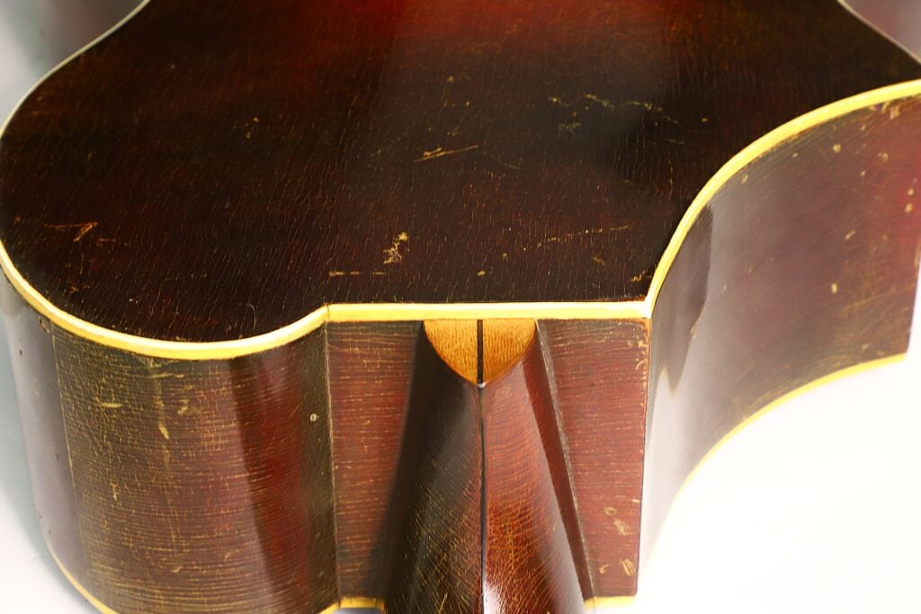 Nahaufnahme der Rückseite und der Zargen einer Epiphone Recording B 1931 Akustikgitarre, die dunkles Holz mit sichtbaren Kratzern und eine helle Einfassung an den Kanten zeigt. Nahaufnahme der Rückseite und der Zargen einer Epiphone Recording B 1931 Akustikgitarre, die dunkles Holz mit sichtbaren Kratzern und eine helle Einfassung an den Kanten zeigt.