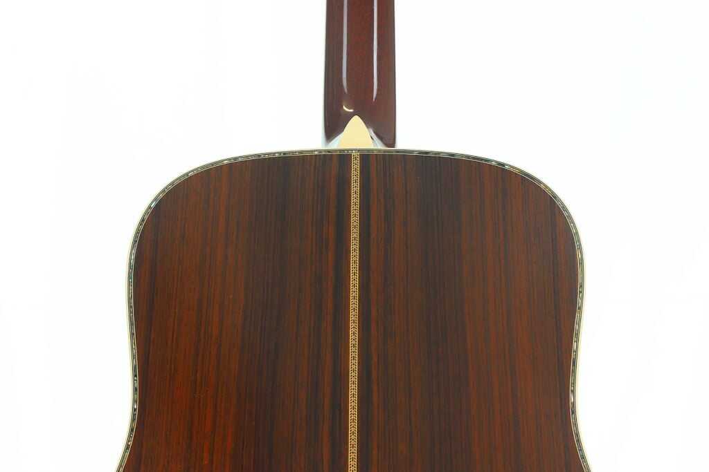 Nahaufnahme der Rückseite einer Martin D-45 Akustikgitarre aus dem Jahr 2009, die die dunkle Holzmaserung und einen vertikal in der Mitte verlaufenden Zierstreifen zeigt. Oben sind der Hals und der Halsfuß der Gitarre teilweise zu sehen. Nahaufnahme der Rückseite einer Martin D-45 Akustikgitarre aus dem Jahr 2009, die die dunkle Holzmaserung und einen vertikal in der Mitte verlaufenden Zierstreifen zeigt. Oben sind der Hals und der Halsfuß der Gitarre teilweise zu sehen.