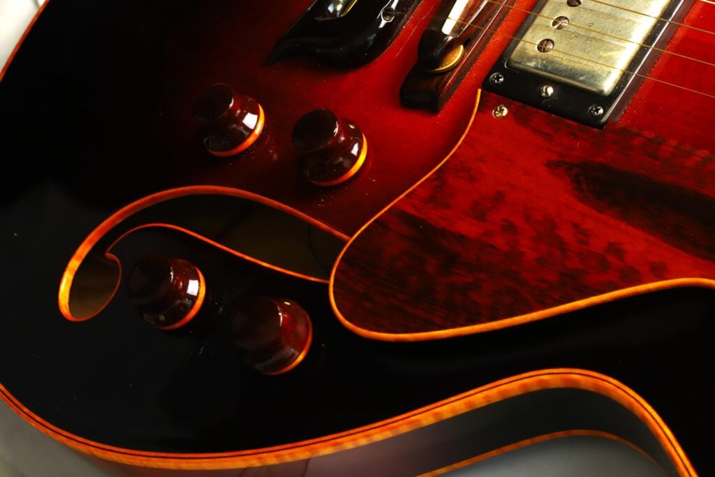 Nahaufnahme des Korpus der DeLaHaye 1995 Elektro-Akustik-Gitarre in Rot und Schwarz, wobei die Regler, ein Teil des Tonabnehmers und die geschwungenen Details der F-Löcher, die durch orangefarbene Zierleisten hervorgehoben werden, im Vordergrund stehen. Nahaufnahme des Korpus der DeLaHaye 1995 Elektro-Akustik-Gitarre in Rot und Schwarz, wobei die Regler, ein Teil des Tonabnehmers und die geschwungenen Details der F-Löcher, die durch orangefarbene Zierleisten hervorgehoben werden, im Vordergrund stehen.