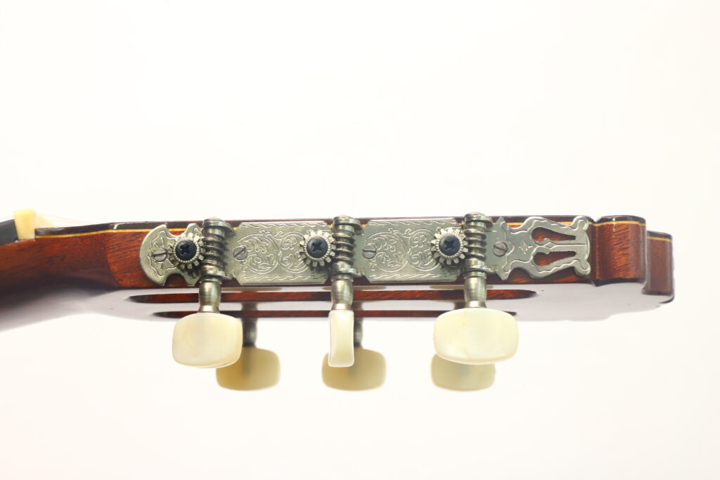Nahaufnahme der Stimmwirbel und der verzierten Metall-Stimmplatte am Kopf einer Flamenco-Gitarre von Casa Arcangel Fernandez aus dem Jahr 1970 vor einem schlichten weißen Hintergrund. Nahaufnahme der Stimmwirbel und der verzierten Metall-Stimmplatte am Kopf einer Flamenco-Gitarre von Casa Arcangel Fernandez aus dem Jahr 1970 vor einem schlichten weißen Hintergrund.