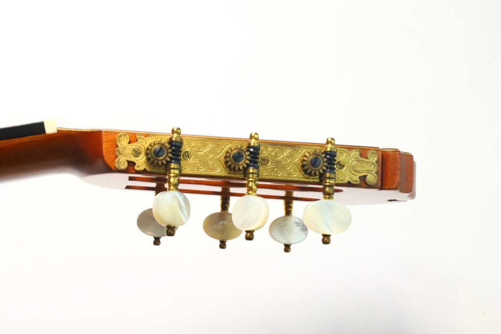 Nahaufnahme der Kopfplatte einer „Masaru Kohno Modell 50 1979“ Klassische Gitarre mit drei kunstvoll gestalteten Stimmwirbeln mit perlweißen Knöpfen und dekorativen goldenen Beschlägen vor einem schlichten weißen Hintergrund. Nahaufnahme der Kopfplatte einer „Masaru Kohno Modell 50 1979“ Klassische Gitarre mit drei kunstvoll gestalteten Stimmwirbeln mit perlweißen Knöpfen und dekorativen goldenen Beschlägen vor einem schlichten weißen Hintergrund.