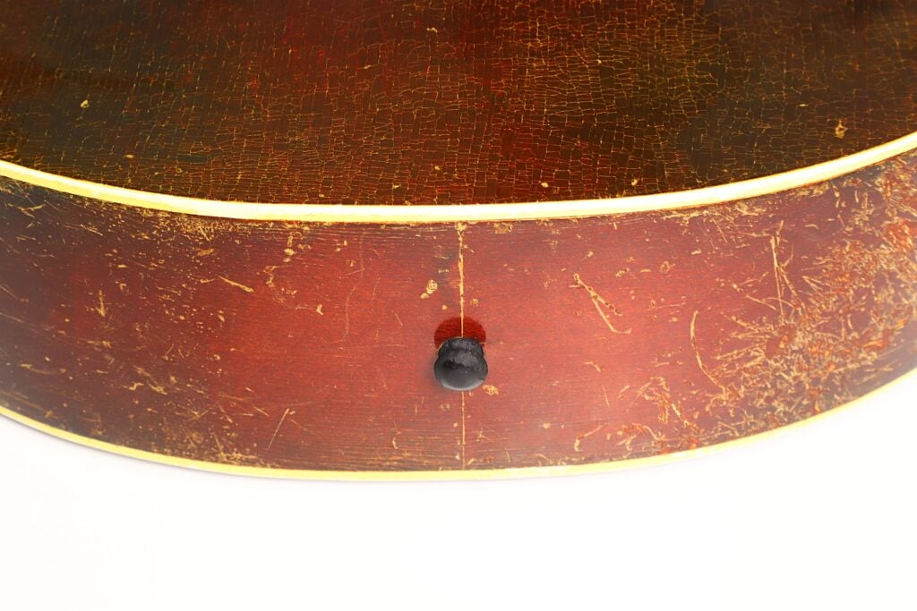 Nahaufnahme der Unterkante einer Epiphone Recording B 1931 Akustikgitarre, die die abgenutzte Vintage-Holzoberfläche mit Kratzern, einen dunklen Gurtknopf in der Mitte der Unterkante sowie eine gelbe Zierleiste entlang der Kante zeigt. Nahaufnahme der Unterkante einer Epiphone Recording B 1931 Akustikgitarre, die die abgenutzte Vintage-Holzoberfläche mit Kratzern, einen dunklen Gurtknopf in der Mitte der Unterkante sowie eine gelbe Zierleiste entlang der Kante zeigt.