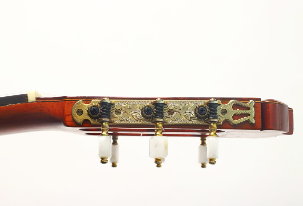 Nahaufnahme des Kopfes einer Ramirez 1a 1985 Klassische Gitarre mit drei Stimmwirbeln, einer verzierten Metallplatte und einer Holzoptik vor einem schlichten weißen Hintergrund. Nahaufnahme des Kopfes einer Ramirez 1a 1985 Klassische Gitarre mit drei Stimmwirbeln, einer verzierten Metallplatte und einer Holzoptik vor einem schlichten weißen Hintergrund.