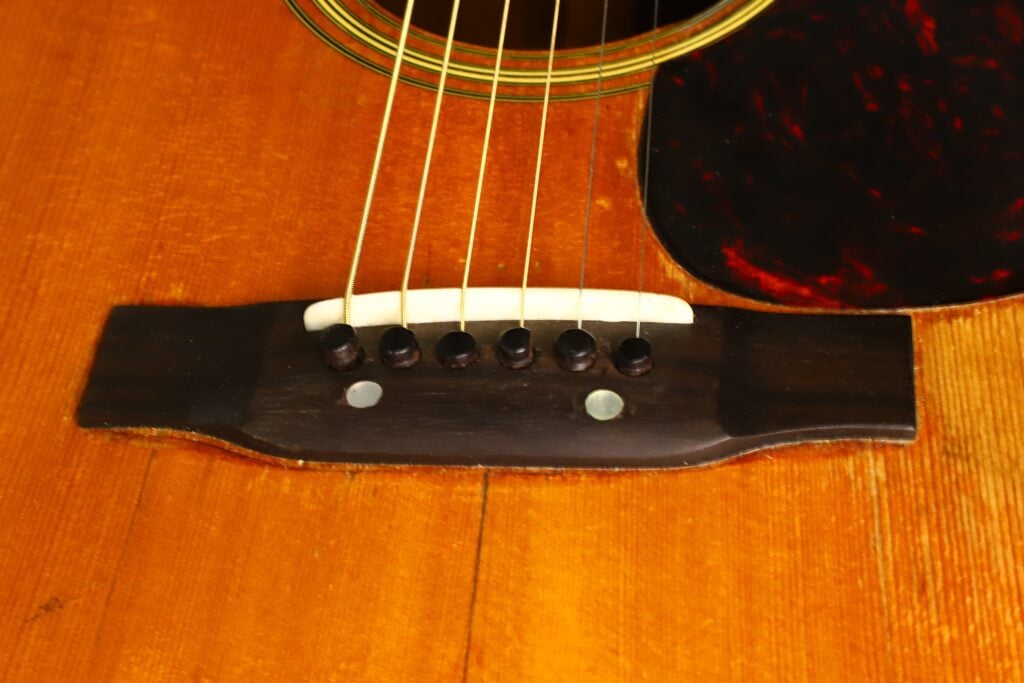Nahaufnahme des Stegs einer Martin D-28 Akustikgitarre aus dem Jahr 1965 mit sechs Saiten, schwarzen Stegstiften und einem weißen Sattel auf dem Holzkorpus. Nahaufnahme des Stegs einer Martin D-28 Akustikgitarre aus dem Jahr 1965 mit sechs Saiten, schwarzen Stegstiften und einem weißen Sattel auf dem Holzkorpus.