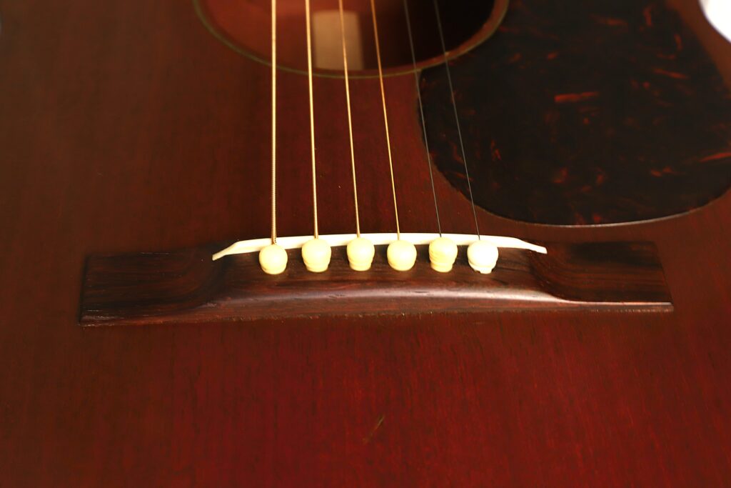 Nahaufnahme des Stegs und des unteren Korpus einer Martin 00-17 Akustikgitarre aus dem Jahr 1941, auf der sechs Saiten zu sehen sind, die mit cremefarbenen Stegstiften auf einer dunklen Holzoberfläche befestigt sind. Nahaufnahme des Stegs und des unteren Korpus einer Martin 00-17 Akustikgitarre aus dem Jahr 1941, auf der sechs Saiten zu sehen sind, die mit cremefarbenen Stegstiften auf einer dunklen Holzoberfläche befestigt sind.